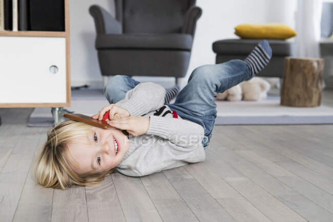 Niño tendido en el suelo, sosteniendo el teléfono inteligente al oído, sonriendo - foto de stock