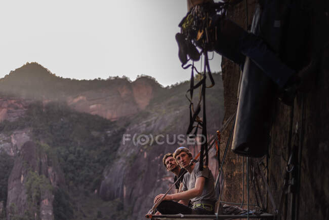 Два скалолаза сидят на портале и смотрят, как друг забирается на скалу рядом с ними, Лимин, провинция Юньнань, Китай — стоковое фото