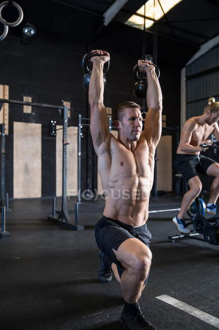 Two men exercising in gymnasium, using kettlebells and air resistance exercise bike — Stock Photo