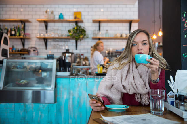 Mujer sentada en la cafetería, sosteniendo el teléfono inteligente, tomando café - foto de stock