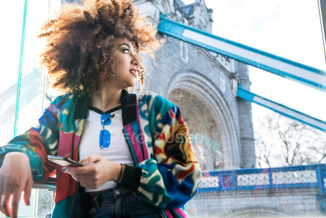 Молода дівчина на відкритому повітрі проведення смартфон, коли хтось дивитися вбік, Тауерський міст у фоновому режимі, Лондон, Великобританія — стокове фото