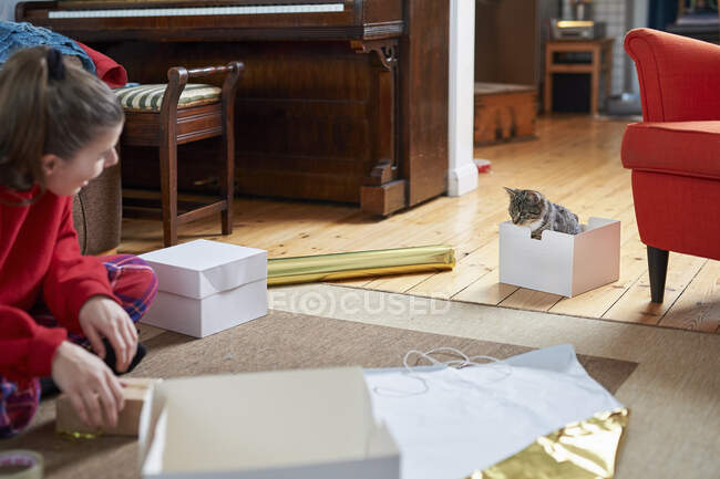 Молодая женщина сидит на полу в гостиной, упаковывая подарки и наблюдая за кошкой — стоковое фото