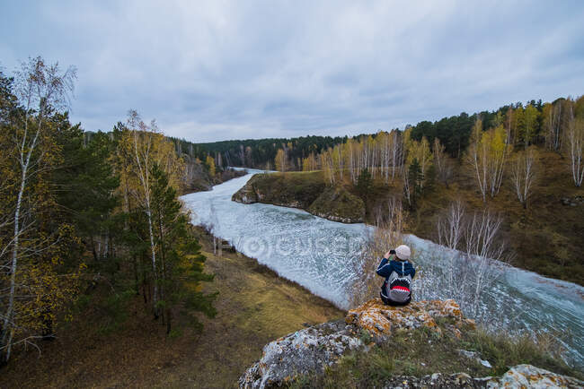 Vista trasera elevada de una joven excursionista fotografiando río desde rocas, Kislokan, Evenk, Rusia - foto de stock