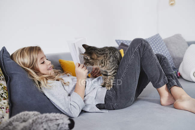 Young girl reading book on sofa with pet cat — Stock Photo