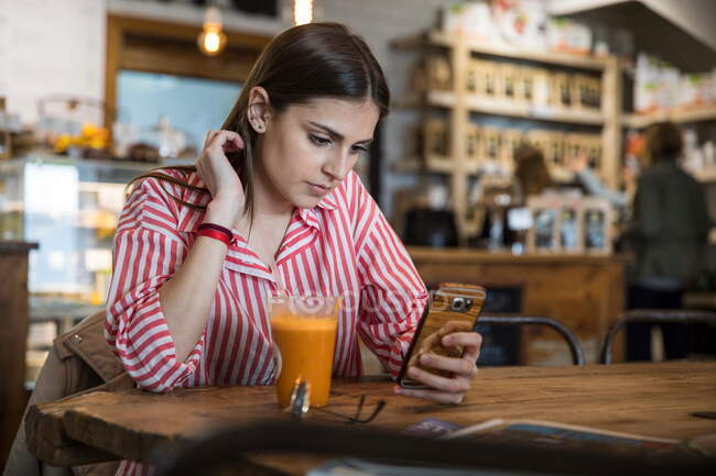 Mujer joven sentada en la cafetería, usando teléfono inteligente, batido en la mesa delante de ella - foto de stock