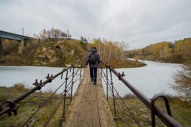Vista trasera del joven caminante masculino caminando por la pasarela del río, Kislokan, Evenk, Rusia - foto de stock