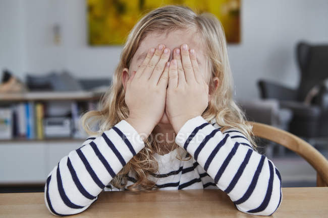 Portrait of young girl at table covering face with hands — Stock Photo