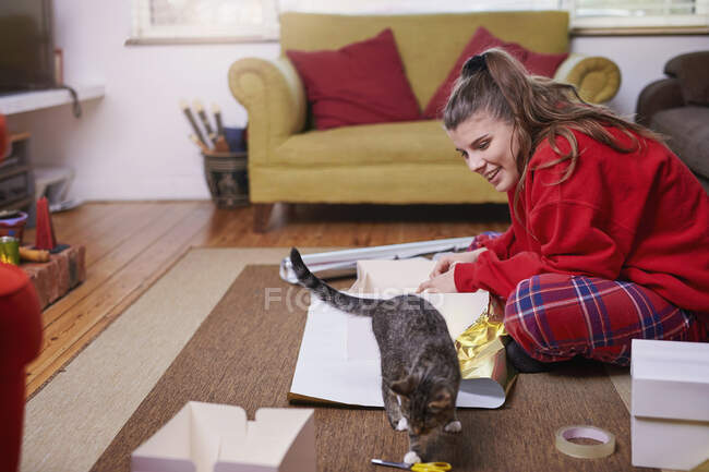 Mujer joven sentada en el piso de la sala de estar envolviendo regalos y mirando gato - foto de stock