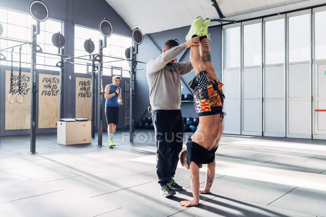 Человек в спортзале помогает другу делать стойку на руках — стоковое фото