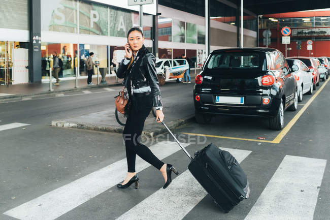 Woman walking with wheeled suitcase and talking on smartphone — Stock Photo
