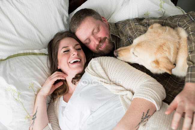 Беременная женщина лежит с партнером на кровати, собака лежит на кровати рядом с ними, возвышенный вид — стоковое фото