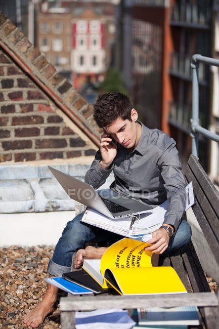 Young man working with laptop and talking on phone on rooftop — Stock Photo