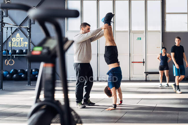 Человек в спортзале помогает другу делать стойку на руках — стоковое фото