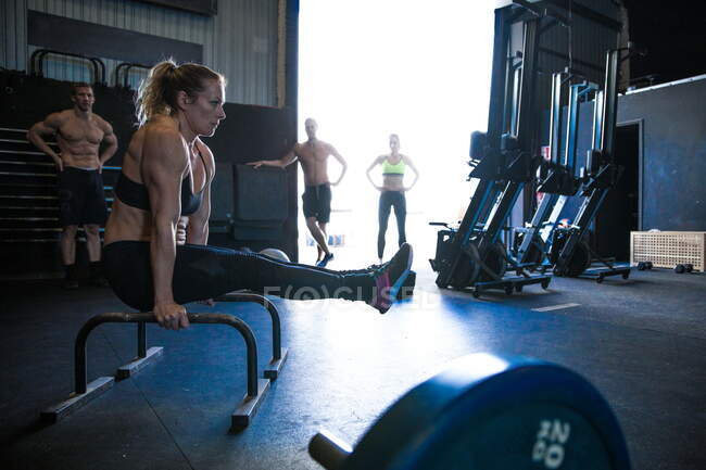 Mujer ejercitándose en el gimnasio, utilizando barras paralelas, en posición L-sit, amigos de fondo, mirando - foto de stock