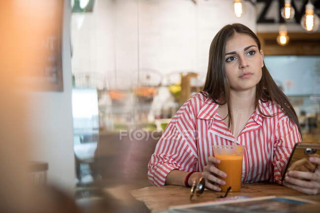 Jovem mulher sentada no café, segurando smartphone, bebendo smoothie — Fotografia de Stock
