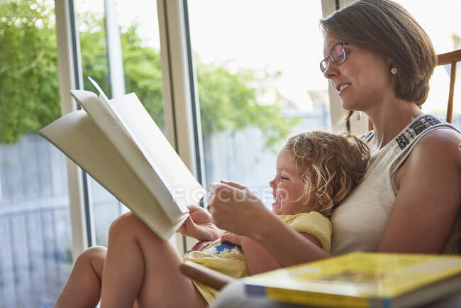 Mother and toddler daughter reading by patio door — Stock Photo
