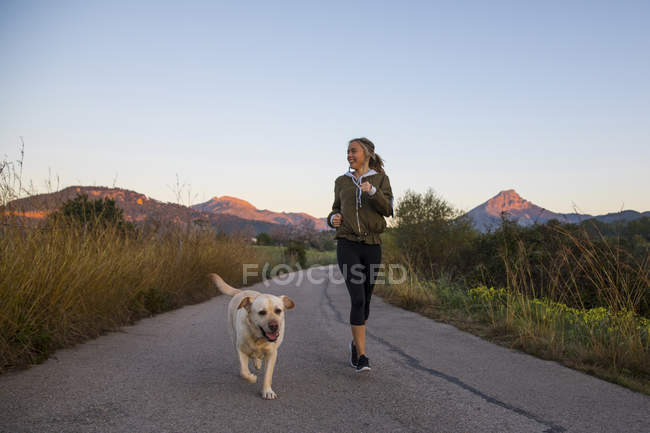 Молода жінка біжить по сільській дорозі з собакою — стокове фото