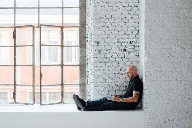 Mann sitzt auf Fensterbank und benutzt Laptop — Stockfoto