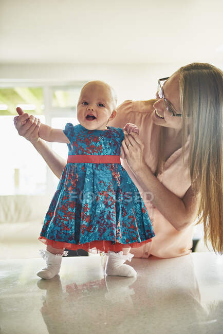 Woman holding hands with baby daughter standing on table, portrait — Stock Photo