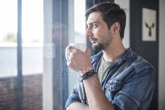 Молодой человек пьет кофе, глядя в окно офиса — стоковое фото