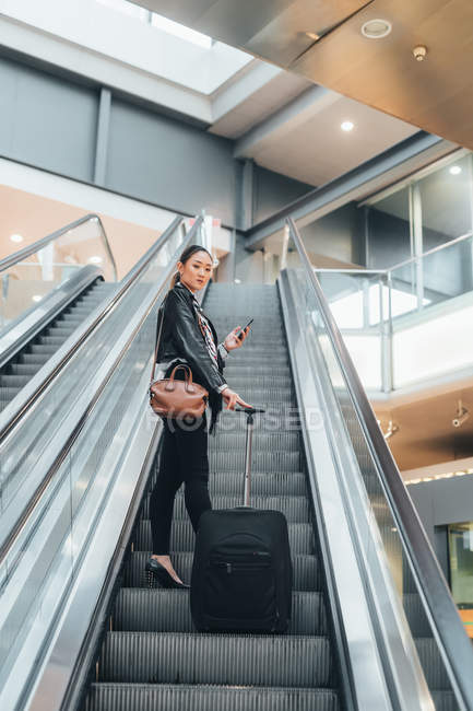 Woman on escalator holding wheeled suitcase and smartphone — Stock Photo
