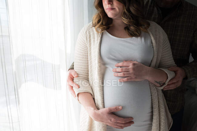 Mujer embarazada tocando el estómago, hombre de pie detrás de ella mostrando afecto, sección media - foto de stock