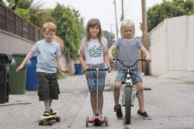 Fille et garçons dans la voie avec scooter, vélo et planche à roulettes — Photo de stock