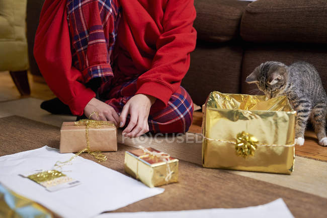 Young woman sitting on living room floor wrapping gifts with curious cat, neck down view — Stock Photo