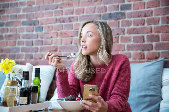 Mujer comer muesli y utilizando smartphone sentado en la cafetería - foto de stock