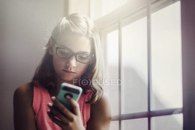 Jeune fille dans des lunettes en utilisant smartphone près de la fenêtre — Photo de stock