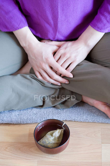 Обрезанное изображение женщины, сидящей в позе лотоса, чашка на полу — стоковое фото