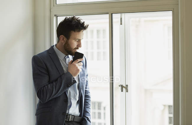 Empresario sosteniendo teléfono inteligente y mirando por la ventana - foto de stock