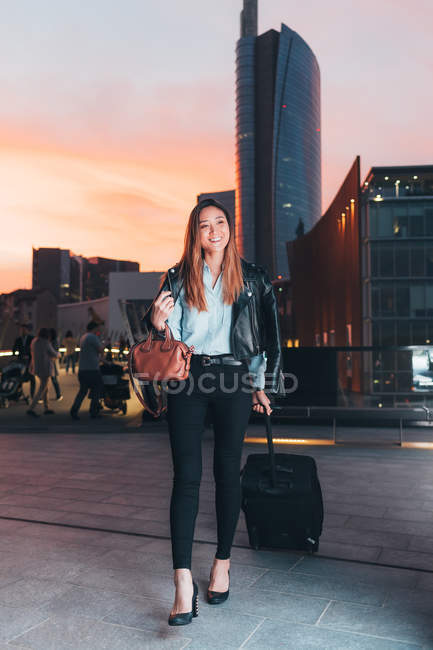 Businesswoman walking outdoors with wheeled suitcase at sunset — Stock Photo