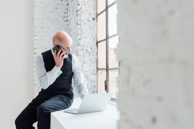 Uomo d'affari sul davanzale della finestra utilizzando laptop e smartphone — Foto stock