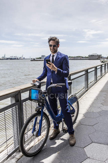 Young businessman on bicycle looking at smartphone along city river waterfront — Stock Photo
