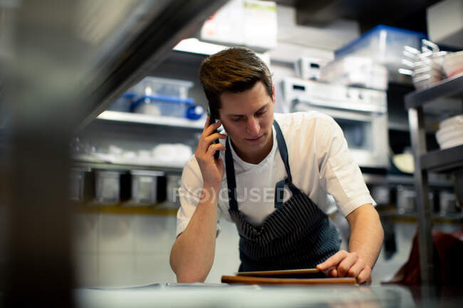 Joven chef masculino leyendo tableta digital y hablando en smartphone en la cocina - foto de stock