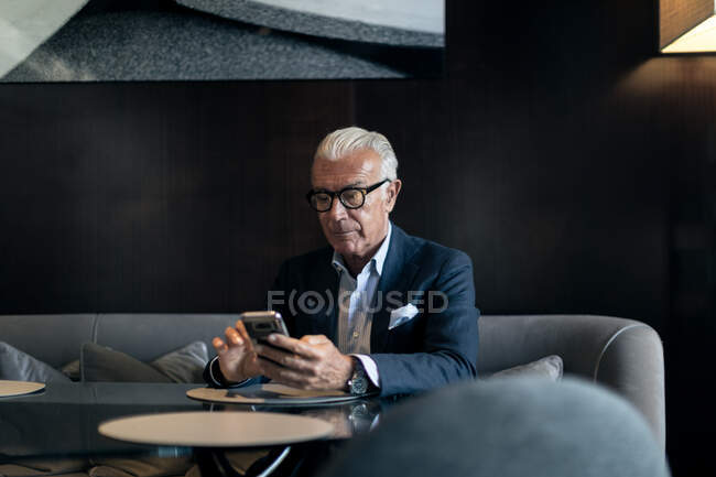 Empresário sênior sentado na mesa do hotel usando tela sensível ao toque do smartphone — Fotografia de Stock
