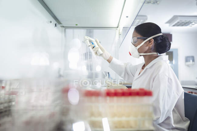 Investigadora científica con máscara y pipeta en el puesto de trabajo del laboratorio de investigación - foto de stock