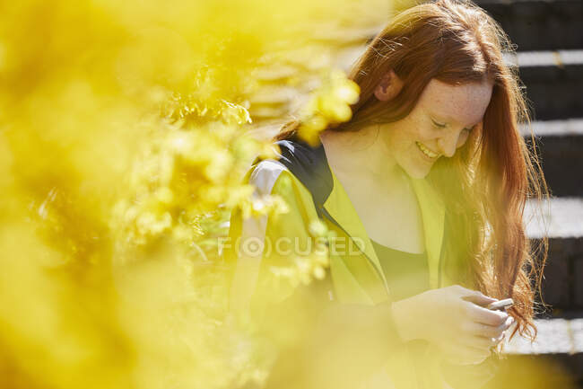 Adolescente assise à l'extérieur sur des marches, vérifiant son téléphone portable, jaune Forsythia au premier plan. — Photo de stock