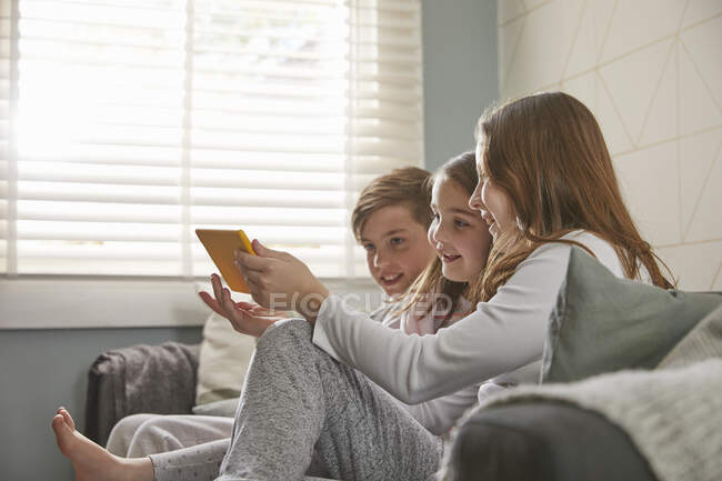 Kindergruppe sitzt im Schlafanzug auf einem Sofa und blickt auf ein digitales Tablet. — Stockfoto