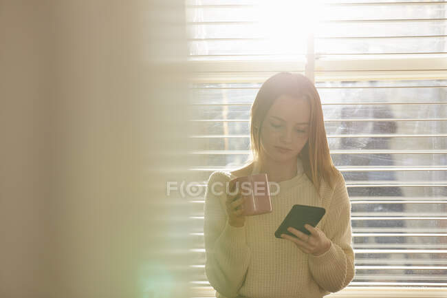 Adolescente de pie frente a la ventana, revisando su teléfono móvil. - foto de stock