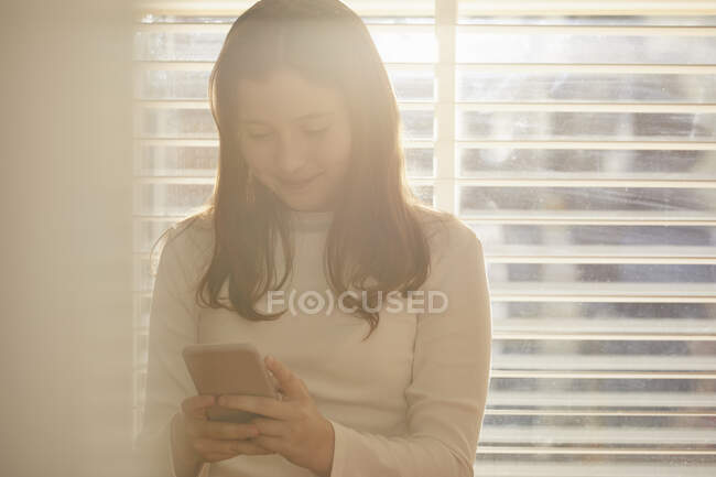 Teenage girl standing in front of window, checking her mobile phone. — Stock Photo