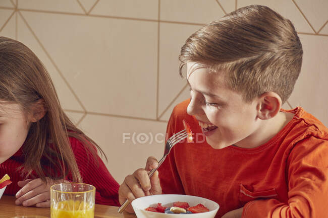 Niño y niña sentados en la mesa de la cocina, desayunando. - foto de stock