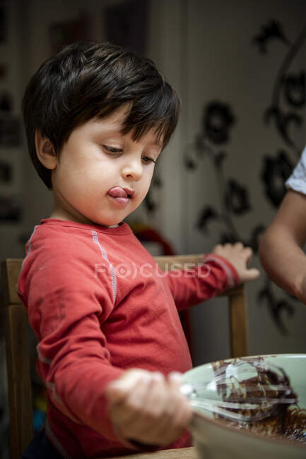 Молодий хлопчик з чорним волоссям сидить за кухонним столом, випікаючи шоколадний торт . — стокове фото