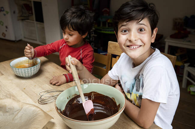 Deux garçons avec les cheveux noirs assis à une table de cuisine, gâteau au chocolat de cuisson. — Photo de stock