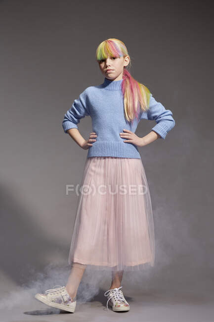 Портрет девушки с длинными красочными окрашенными волосами и окрашенной бахромой в голубой свитер и розовую юбку-пачку, смотрящей в камеру, на сером фоне, руки на бедрах — стоковое фото