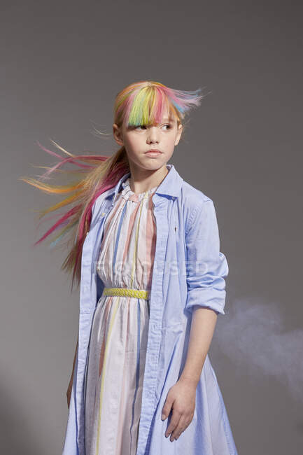 Portrait of girl with long colorful hair and dyed fringe wearing white dress and pale blue coat, looking away at grey background — Stock Photo
