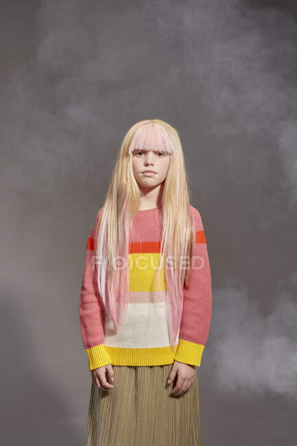 Portrait of girl with long blonde hair wearing yellow and red stripe top and khaki skirt, looking at camera, on grey background — Stock Photo