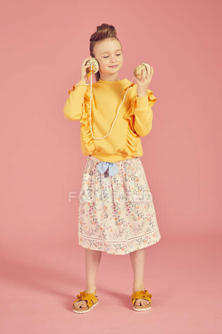 Portrait of brunette girl wearing yellow top and skirt with floral pattern holding sea shell phone, on pink background — Stock Photo