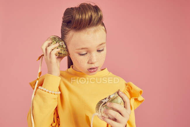 Brunette girl wearing yellow top, holding sea shell phone, on pink background, looking on shell — Stock Photo